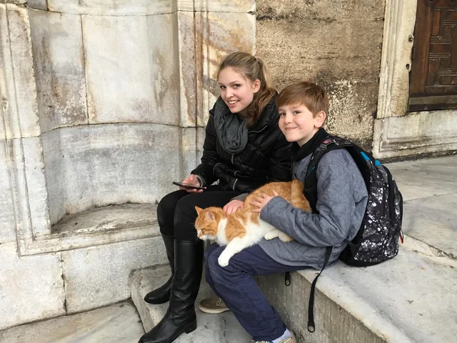 Two young travellers pausing with a cat beside historic stonework.